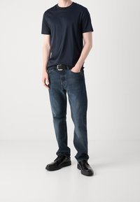 Man som står med ena handen i fickan iförd en marinblå T-shirt, mörkblå jeans med svart bälte och svarta läderskor på en enhetlig bakgrund.