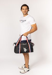 Sac de sport noir avec des rayures rouges et bleues, affichant un logo à l'avant. Le matériau semble lisse. Fermeture zippée et poignées solides.