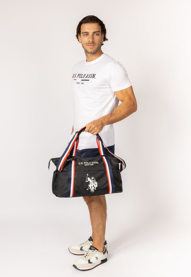 Sac de sport noir avec des rayures rouges et bleues, affichant un logo à l'avant. Le matériau semble lisse. Fermeture zippée et poignées solides.