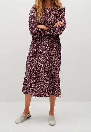 Femme debout, bras croisés, portant une robe midi à manches longues à motif floral bordeaux et des chaussures blanches tissées à enfiler sur fond uni.