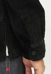 Black corduroy jacket with vertical ribbed texture, featuring a buttoned cuff and side slit, worn with dark denim showcasing red stitching.