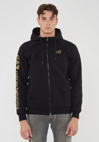 Felpa nera con zip, cappuccio regolabile, due tasche frontali e logo giallo sulla manica sinistra e sul petto. Materiale misto cotone.