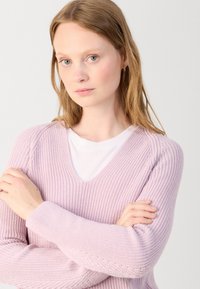 Pulover tricotat de culoare mov deschis, cu decolteu în V, țesătură texturată și mâneci lungi, purtat peste o cămașă albă. Brațele încrucișate în față.