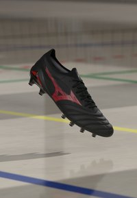 Schwarzer und roter Fußballschuh mit Stollen, der über einem Hallenboden schwebt, der mit farbigen Linien markiert ist.