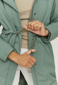 Light green padded jacket with a drawstring closure, featuring a stand-up collar and zipper. Hands adjust the drawstring at the waist.