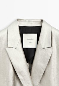Lichtgrijze linnen blazer met piekrokken, voorzien van een zwarte voering en een label met de tekst "Massimo Dutti Studio" en onderhoudsinstructies.