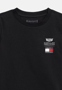 Μαύρο μπλουζάκι crewneck με λευκό λογότυπο της ομάδας Cadillac Formula 1 και λογότυπο με τη σημαία Tommy Hilfiger στο πάνω αριστερό μέρος του στήθους.