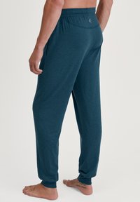 Teal loungebroek met een ontspannen pasvorm, elastische tailleband, zijzakken en ribgebreide enkelboorden. De stof lijkt zacht en lichtgewicht.