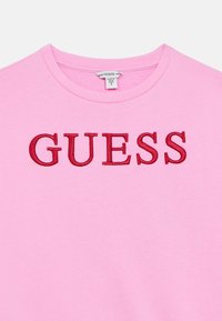 Ροζ φούτερ με ριμπ λαιμόκοψη. Διαθέτει μεγάλο, κόκκινο κεντητό λογότυπο "GUESS" στο στήθος. Μαλακό βαμβακερό υλικό.