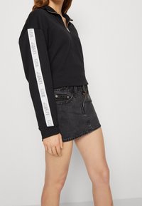 Černá mikina se zipem do čtvrtiny a bílými detaily loga na rukávech, kombinovaná s černou džínovou sukní s vysokým pasem.