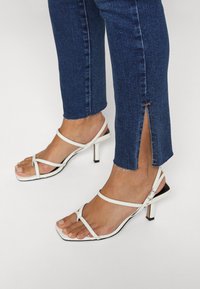 Vita remmar med klacksandaler med fyrkantig tå och svart accentklack, kombinerat med mörka jeans i denim med rå fåll och sidöppning.