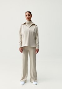 Beige sportos szett, hosszú ujjú pólóval, amelynek gallérja van, és széles szárú nadrággal, puha, sima anyagból készült, fehér cipővel párosítva.