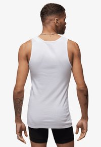 White cotton tank top with a ribbed texture, a round neckline, and wide shoulder straps, worn over black shorts. Back view shows a fitted design.