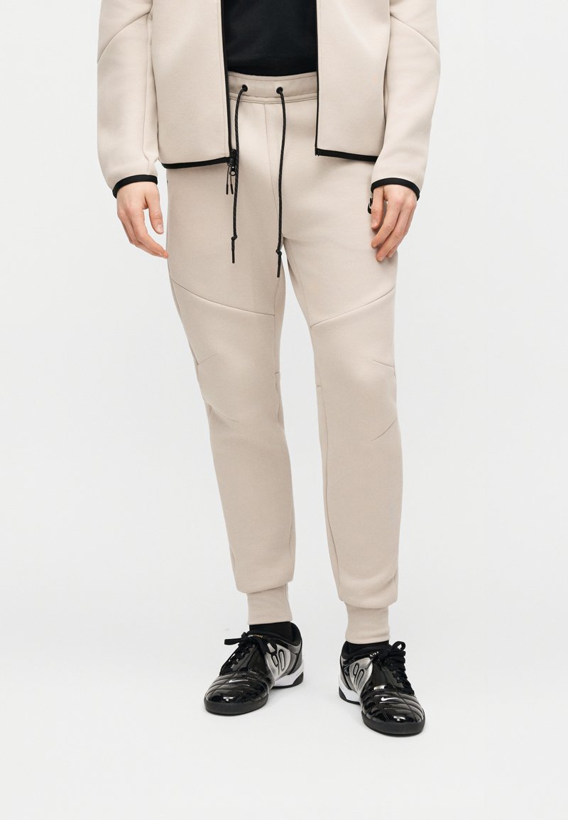 Pantalones jogger beige con bolsillos con cremallera, cordón negro, combinados con zapatillas negras y plateadas con cordones, y una chaqueta beige sobre una camiseta negra.