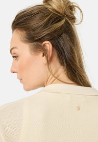Pull en maille beige clair présentant un tissu texturé, un col et un petit bouton accent au dos du cou.
