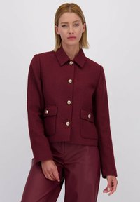 Vrouw in een gestructureerde bordeauxrode jas met gouden knopen en bijpassende bordeauxrode broek, staand tegen een effen lichte achtergrond.