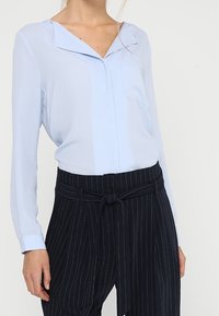 Ljusblå blus med V-ringning och långa ärmar, med en bröstficka. Tillsammans med mörka randiga byxor och ett knutet midjeband.