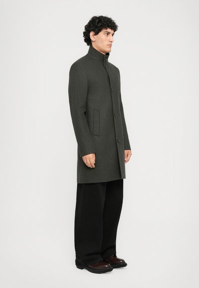 Manteau en mélange de laine grise avec col haut, fermetures à boutons, poches latérales et coupe ajustée, associé à un pantalon ample noir et des chaussures marron.