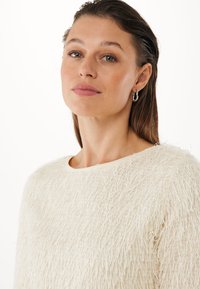 Maglione beige testurizzato con scollatura rotonda, maniche lunghe e una finitura morbida e pelosa. Orecchini a cerchio in argento completano il look.