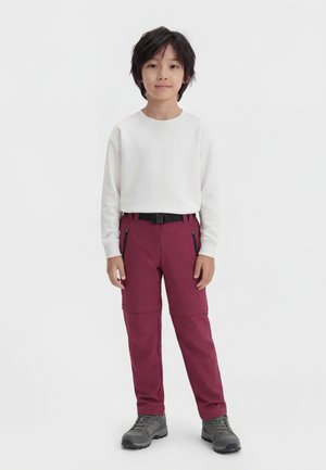 Dieťa s čiernymi vlasmi má na sebe bielu dlhú rukávovú košeľu, vínové nohavice na zips s čiernym opaskom a šedé turistické topánky, stojí na bielom pozadí.