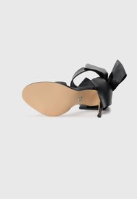 Black high-heeled sandal with a wide bow accent, open design, and a smooth beige sole. The heel is thin and pointed.