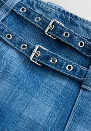 Indumento in denim con un motivo a quadri blu, dotato di hardware argentato e numerosi grommets per cinghie regolabili. Testura del tessuto classica.