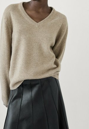 Personne portant un pull beige en V rentré dans une jupe en cuir noire taille haute à coupe trapèze, sur un fond uni.