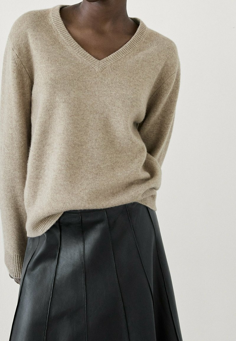 Personne portant un pull beige en V rentré dans une jupe en cuir noire taille haute à coupe trapèze, sur un fond uni.