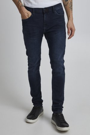 Une personne tatouée porte un jean foncé ajusté et des baskets noires, une main dans une poche avant.