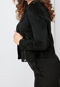 Veste en suède noir avec des zips argentés, au design ajusté, et des détails subtils sur les épaules et les poignets. Texture lisse avec une silhouette élégante.