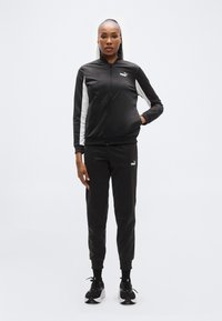 Μαύρο αθλητικό σετ φόρμας με ζακέτα με φερμουάρ που διαθέτει λευκές πλαϊνές πινελιές, συνδυασμένο με αντίστοιχο παντελόνι φόρμας. Φοριέται με μαύρα και λευκά αθλητικά παπούτσια.