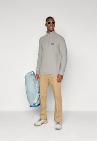 Γκρι φούτερ από φλίς με φερμουάρ στο ένα τέταρτο, καφέ παντελόνι και ανοιχτό μπλε σακίδιο. Λευκά αθλητικά παπούτσια και γυαλιά ηλίου ολοκληρώνουν την εμφάνιση.