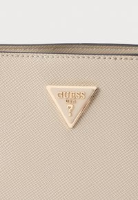 Matière synthétique texturée beige avec un logo triangulaire doré portant l'inscription "GUESS USA" et un point d'interrogation au centre.