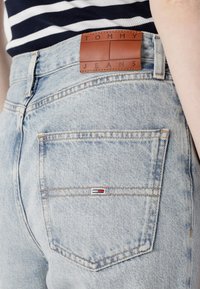 Heledad sinine denim püksid, mille vöökoht on märgistatud nahaplaadi külge "TOMMY JEANS" ja millel on väike logo tagataskus.