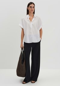 Camicia bianca oversize con bottoni, maniche arrotolate, abbinata a pantaloni neri a gamba larga e una borsa a spalla intrecciata scura. Tessuto dalla texture morbida.