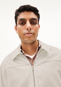 Giacca beige con zip e colletto sopra una camicia a quadri marrone chiaro. Il tessuto sembra morbido con una superficie liscia.