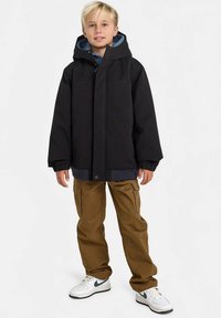 Blond deček stoji, oblečen v črno kapuco jakno, rjave cargo hlače in bele superge z temno zelenimi swoosh logotipi.