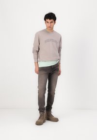 Felpa grigia chiara con logo "BELSTAFF", sovrapposta a una maglietta verde menta. Abbinata a jeans grigi scuri e stivali chiari con lacci.