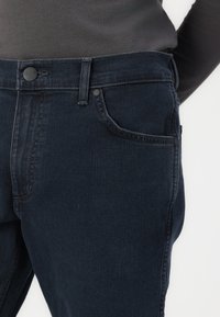Jeans azul oscuro de mezclilla, con un bolsillo frontal, cierre de botón y un diseño simple con una ligera textura.