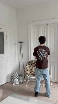 Braunes T-Shirt mit grafischem Druck auf der Rückseite, kombiniert mit hellblauen Jeans und schwarzen Schuhen. Weiße moderne Lampe und dekorativer Stuhl im Hintergrund.