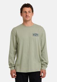 Uomo con capelli corti e scuri, che indossa una camicia a maniche lunghe di colore verde chiaro con un piccolo logo grafico sul lato sinistro del petto, in piedi davanti a uno sfondo bianco.