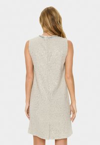 Ärmelloses, hellbeiges Kleid mit einer strukturierten, glitzernden Oberfläche und einem mit Silber verzierten Ausschnitt. Mittellang bis zur Oberschenkellänge mit geradem Schnitt und Schlitz auf der Rückseite.