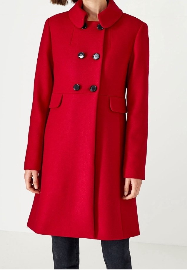 Manteau rouge croisé avec des boutons noirs, des poches à rabat et un col arrondi porté sur un pantalon sombre.