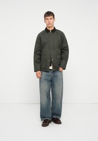 Donkergroen gewatteerde jas met een bruine corduroy kraag, twee voorzakken, gecombineerd met lichtblauwe wijde jeans en bruine schoenen.