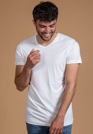 Weißes V-Ausschnitt-T-Shirt aus leichtem Stoff, figurbetontes Design, kurze Ärmel, getragen mit blauen Jeans vor einem neutralen Hintergrund.