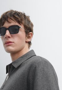 Grau zipfeljacke mit Kragen, kombiniert mit schwarzen quadratischen Sonnenbrillen. Der Hintergrund ist schlicht weiß, was die Kleidung und Accessoires betont.