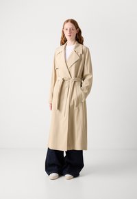 Beiger Trenchcoat mit breiten Revers, tailliertem Gürtel und Seitentaschen, kombiniert mit weiten dunklen Jeans und weißen Sneakers. Glatte Textur.