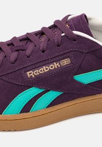 Fialový semišový teniska s tyrkysovými akcenty, zlatým logem "Reebok" a texturovanou gumovou podrážkou barvy přírodní gumy. Tkaničky ladí s barvou a designem boty.