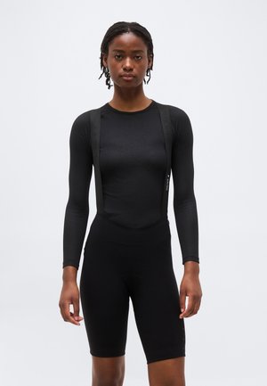 Kobieta z warkoczem, ubrana w dopasowaną czarną bluzkę z długim rękawem oraz czarne, dopasowane szorty kolarskie z szelkami, stojąca na jednolitym jasnym tle.