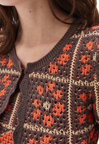 Primer plano de un cardigan marrón de punto con patrones florales geométricos en naranja y crema en la parte superior del pecho y el cuello de una persona.